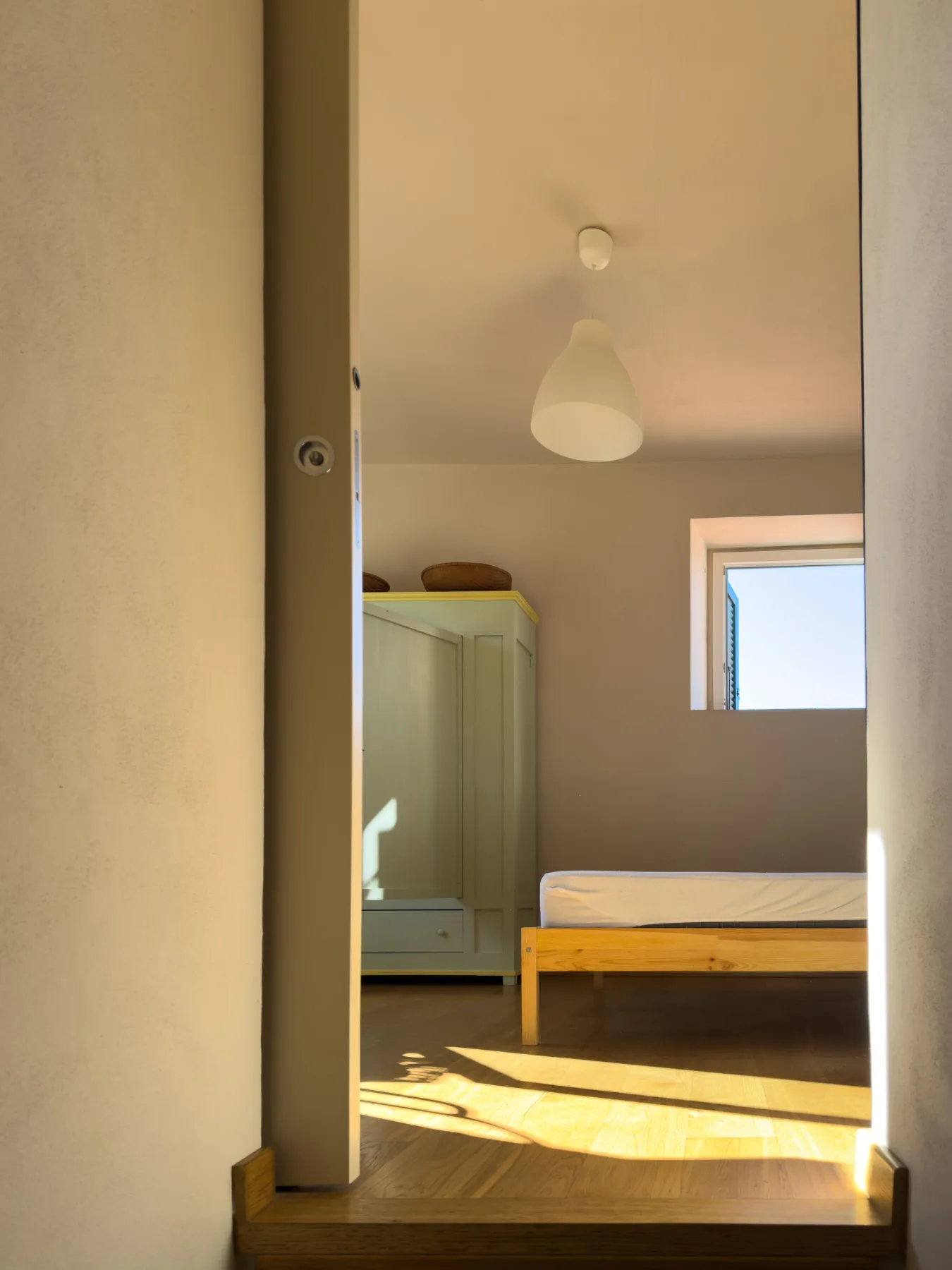 Bedroom viewed through an open doorway, afternoon light across a wood floor.